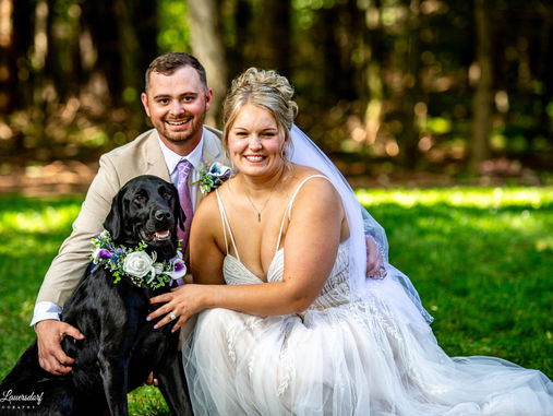 a bride and groom kneel down next to their black lab with a flower decorated collar.