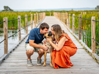 A man in a blue shirt, and a woman in an orange dress lean down to kiss their dog on the head while on the floating boardwalk path in the Horicon marsh