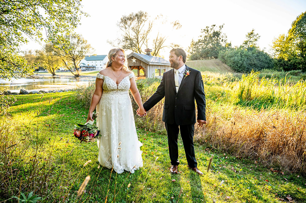 A bride and groom hold hands, smiling in a sunlit field. The bride wears a white gown and holds flowers. A rustic building and trees are in the background.