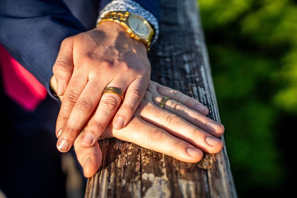 Hands with rings rest on a wooden rail, one golden watch visible. Sunlight creates a warm, intimate feel against a blurred green background.