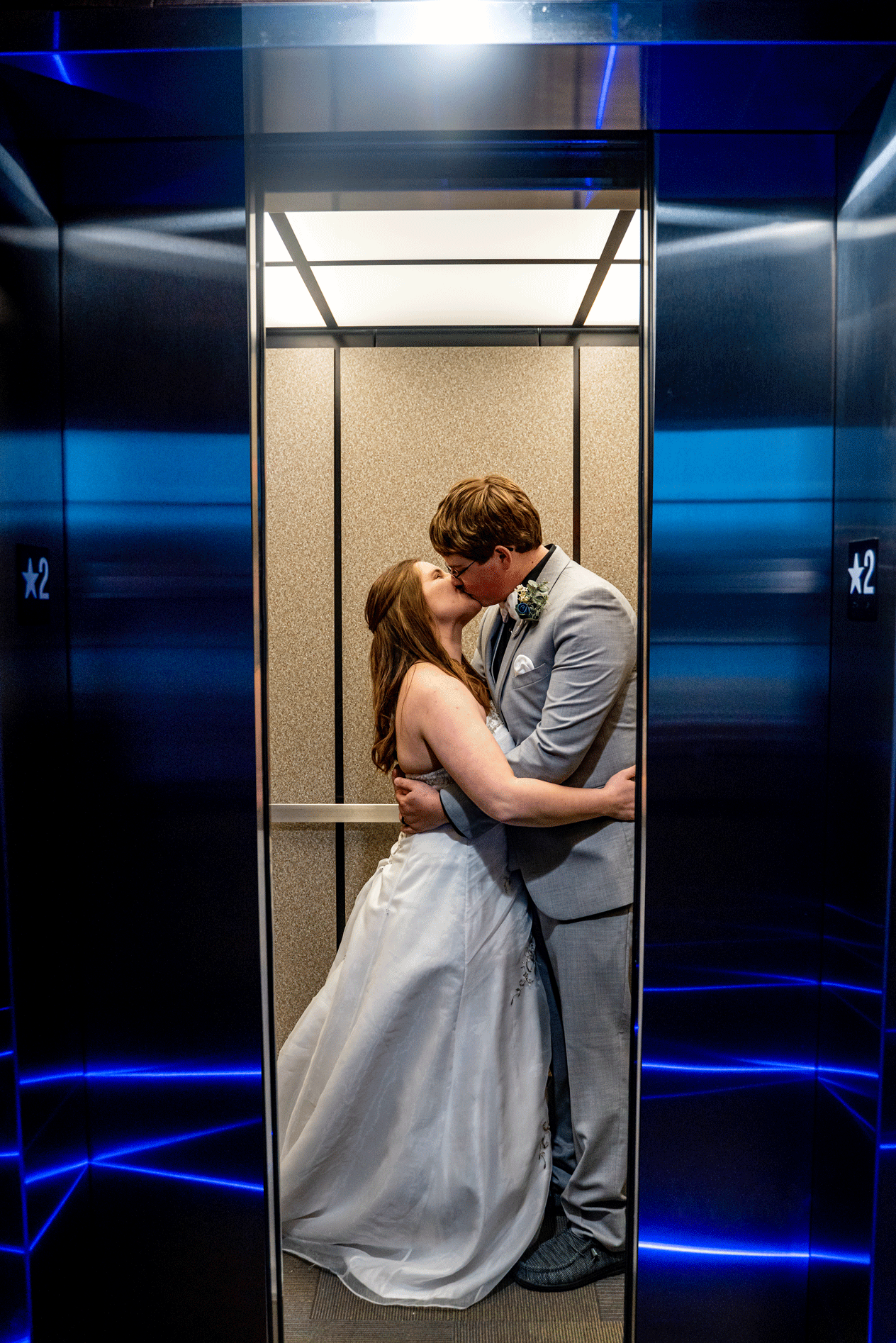an elevator door closes as a bride and groom kiss. Blue lights reflect off the metal elevator