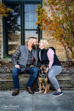 a couple leans in for a kiss while sitting in front of a brick building with their dog