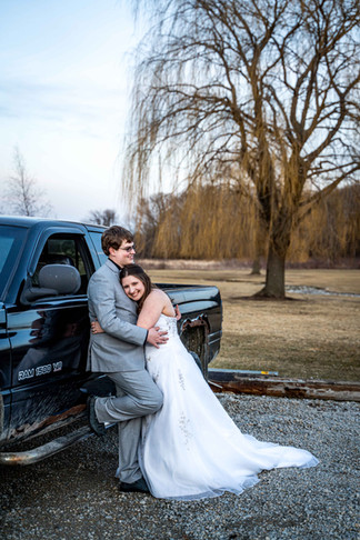 A bride in white floor length dress rests her head on groom's chest. Groom is in a grey suit leaning up against black pick up truck. a willow tree and sky in the background