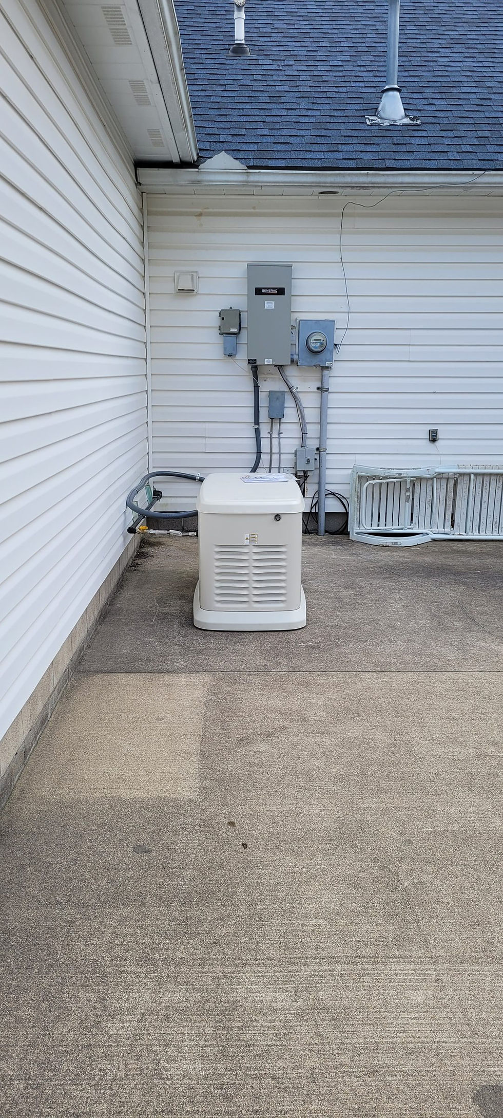 Finished standby generator on a concrete patio with a Generac transfer switch and meter on the wall, seen from a distance.