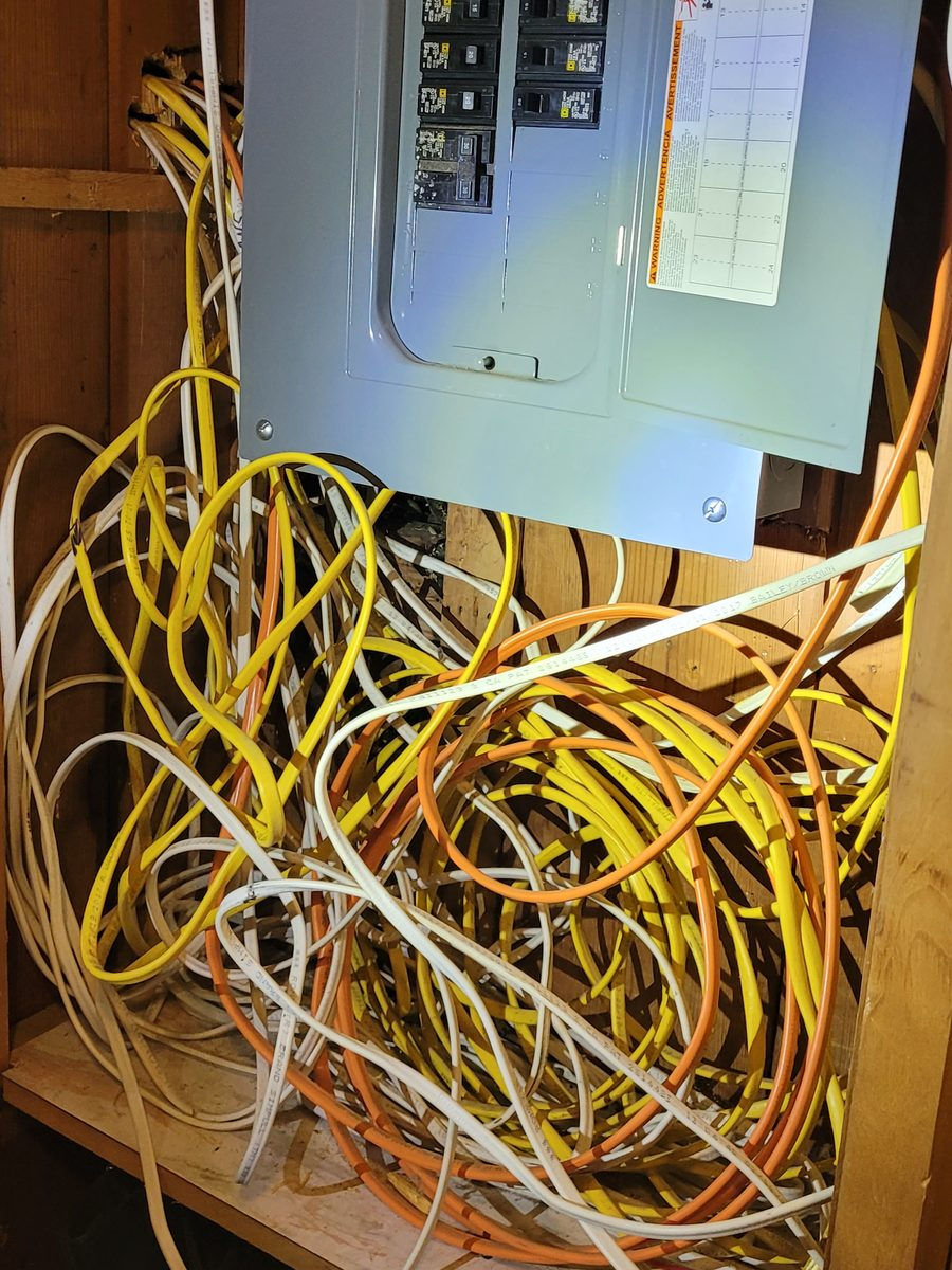 Breaker panel on a wood wall with a large tangle of white, yellow and orange cable below it