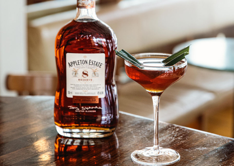 Un cocktail dans un verre à martini garni d'une feuille verte, placé à côté d'une bouteille de rhum Appleton Estate 8 ans de réserve sur une table en bois avec un éclairage doux depuis une fenêtre. Photo prise par Folly Arts & Medias, photographe de cocktails.