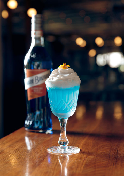 A whimsically presented blue cocktail topped with whipped cream and an orange zest, accompanied by a bottle of Marie Brizard Curacao Blue on a wooden bar. Picture taken by Folly Arts & Medias, cocktail photographer.