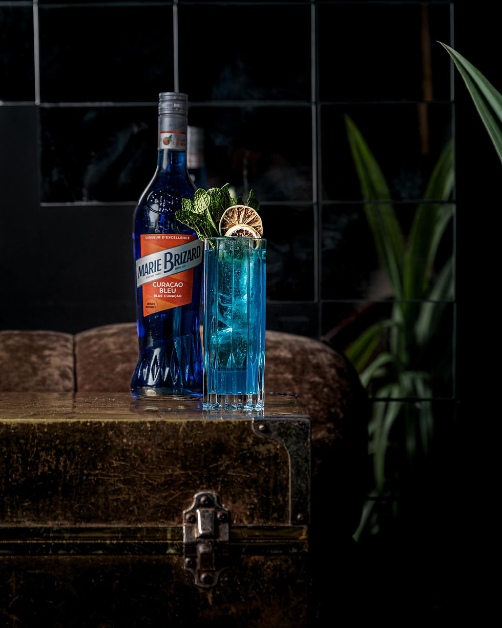 A blue cocktail resting on a box, next to a bottle of Marie Brizard Blue Curaçao