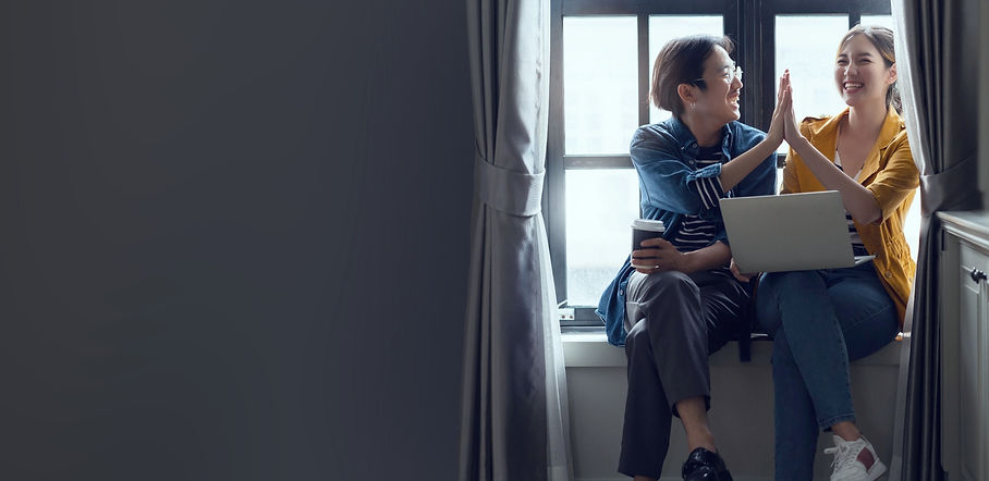 A girl and guy friends high-fiving whilst sitting against a window. Both are laughing, one has a coffee cup and the other has laptop on her knee.