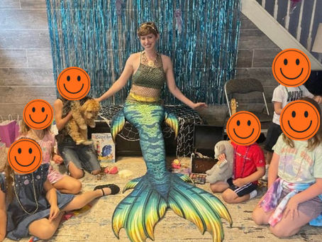 mermaid liv at a mermaid gig with children, wearing fin fun elite mermaid tail. there is a blue tinsel backdrop and mermaid liv is sitting on a bench with fishnet. On the floor there is a treasure chest and some toys and props are scattered around. the book Beachy and Me is sitting against the bench. one little girl is holding a dog.