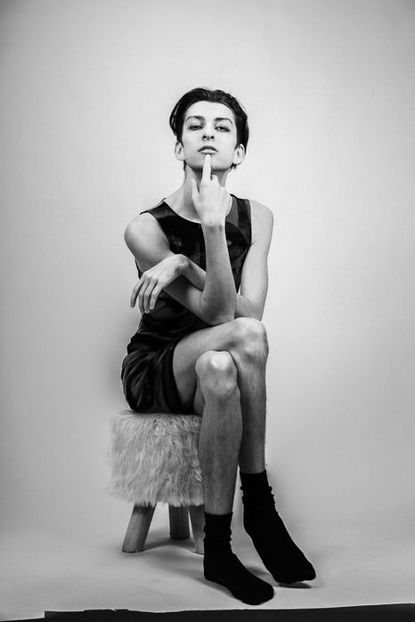 A young man in a dress sitting on a fluffy stool with crossed legs, striking a sultry pose, against a white background, rendered in high contrast black and white.