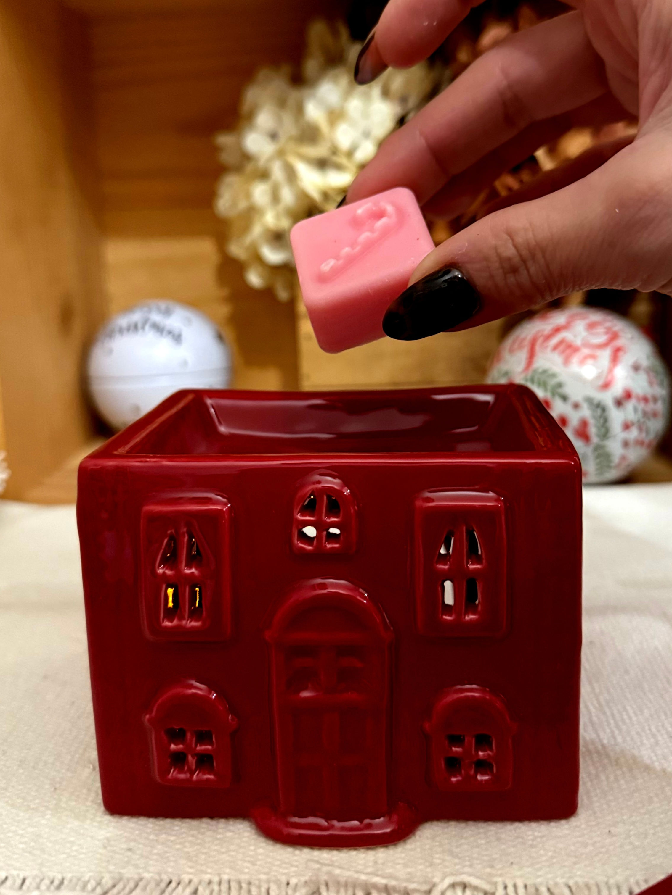 Maison rouge de Noël en céramique, main plaçant un fondant parfumé rose sur la coupelle.