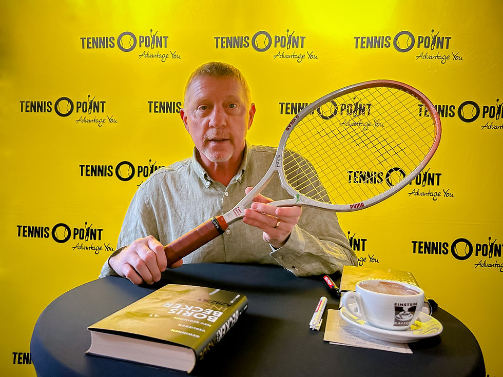 Former professional tennis player Boris Becker holding a Puma G Vilas Tennis racquet, signed by Boris Becker, Manager Ion Tiriac, and coach Günther Bosch, sitting at a chair in front of a yellow Tennis Point commercial wall.