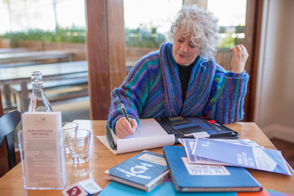 Dr Annetta Mallon is sitting at a small square table with her back to a window. The table is covered in papers and books relating to advance planning.