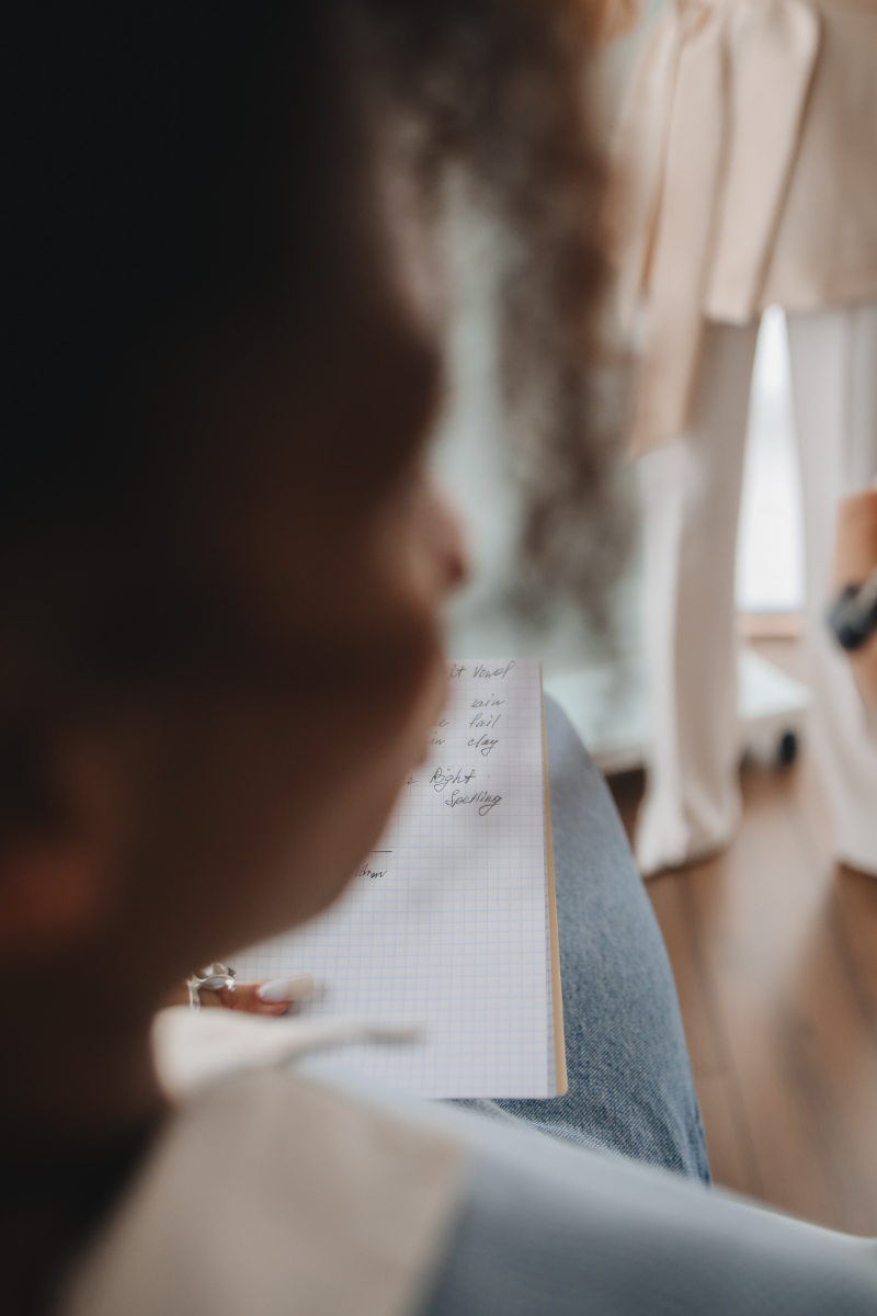 A woman with dark skin in blurred side profile sits with a notebook on her lap. The photo is taken from over the woman's right shoulder, and she is wearing jeans.