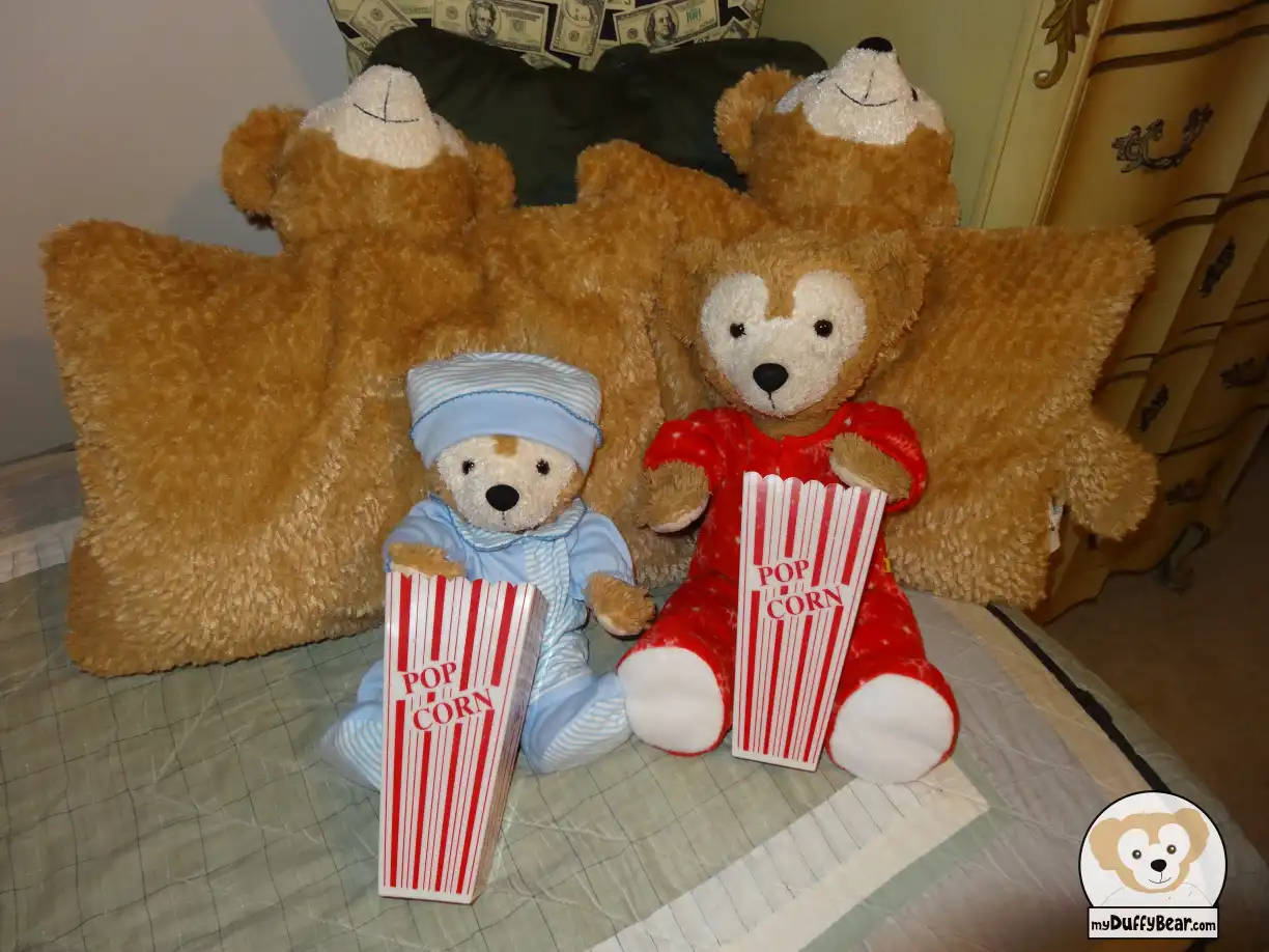 Duffy and Little Joe are dressed in their pajamas, each of them with a Duffy the Disney Bear pillow pet behind them as they have a paw in a special red with white stripes plastic popcorn container that mimics a paper popcorn bag