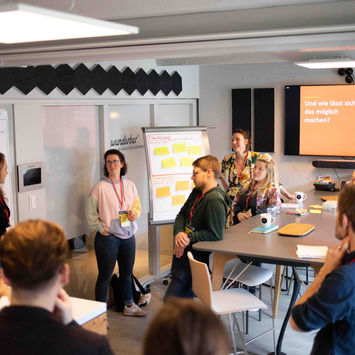 Teilnehmer:innen eines Workshops präsentieren ihre Ideen vor Flipchart und Bildschirm in einem modernen Seminarraum.