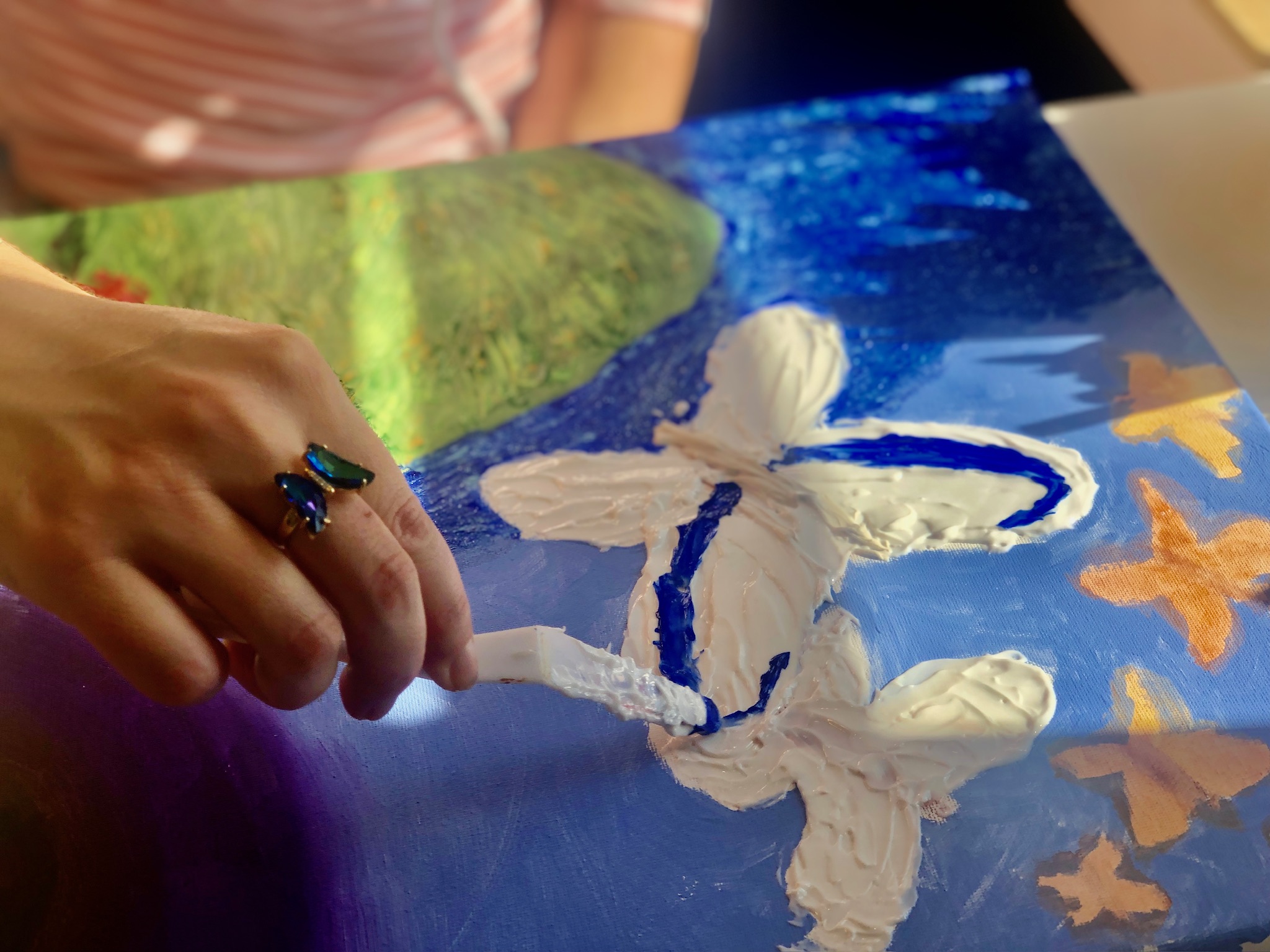 A hand wearing a butterfly ring paints a white butterfly onto a blue and gold canvas