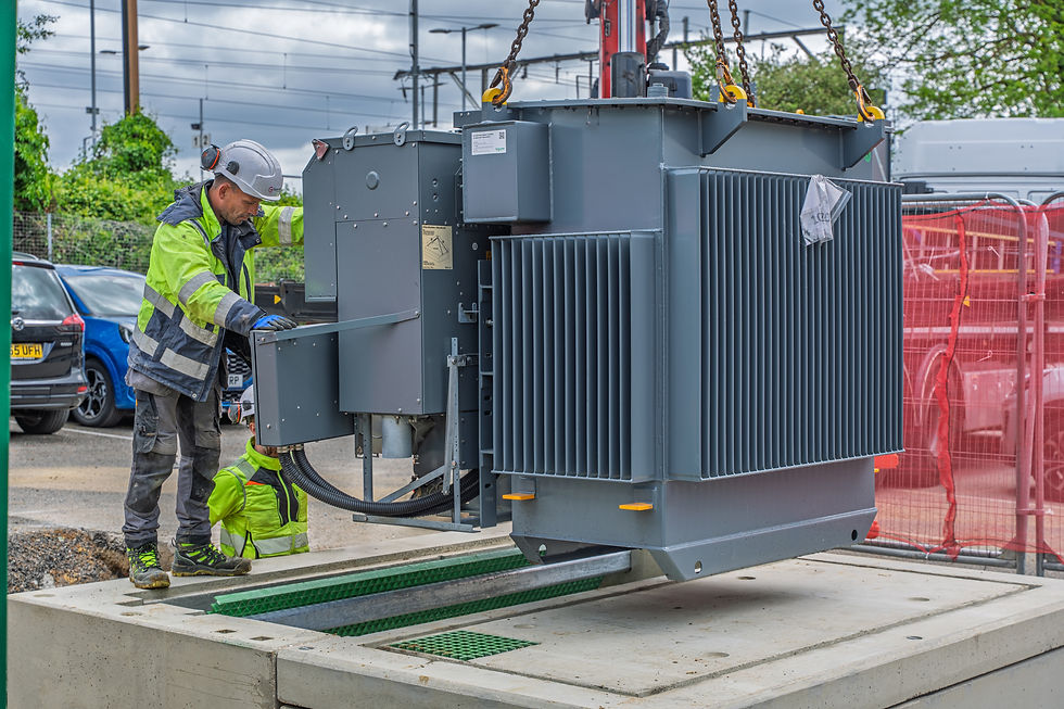 Workers in hi-vis gear install a large grey substation with crane chains at a roadside utility site, under cloudy skies.