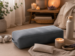 A serene meditation space featuring a yoga bolster on a mat, a cozy folded blanket, a lavender eye pillow, candles, soft lighting, eucalyptus, headphones, and a water bottle. The calming neutral décor creates a peaceful environment for meditation, mindfulness, and evening relaxation.