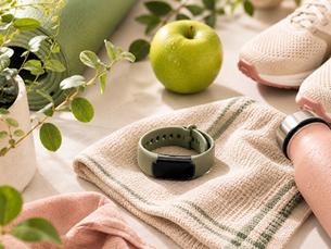 Fitness gear setup with a green smartwatch on a towel, green apple, pink sneakers, and water bottle on a light background, surrounded by plants.