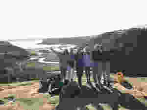 six girls all smile on a hill top, arms behind each others back, with the girls on both ends with their arms in the air. The background shows hills and a river leading to a beach.