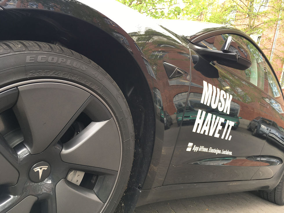 Close-up of a black Tesla with a large wheel and side decal reading MUSK HAVE IT., parked on a tree-lined street.