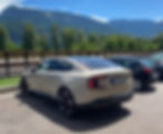 Beige Volvo ES90 sedan parked beside a black hatchback, with green mountains and a blue sky in the background on a sunny day.