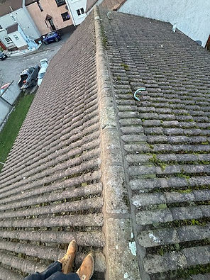 A person wearing boots stands on the peak of a mossy, weathered tiled roof, overlooking a quiet residential street with parked cars below