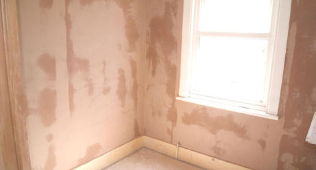 A small, unfinished room with freshly plastered walls and a single window letting in bright natural light