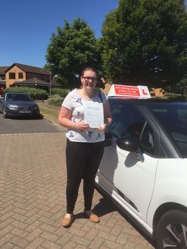 A person stands smiling beside a white car with an "L" sign on top, holding a certificate.