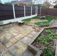 A backyard with rectangular turf bordered by light-colored tiles, a raised wooden garden bed, a wooden shed, and a privacy screen, adjacent to a brick and stucco house