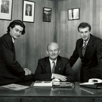 Three men in suits pose in an office with wood-panelled walls and framed photos