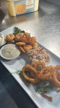 A white rectangular plate with crispy calamari, sautéed shrimp, shredded crab, and two dips. The setting is a stainless steel kitchen surface.