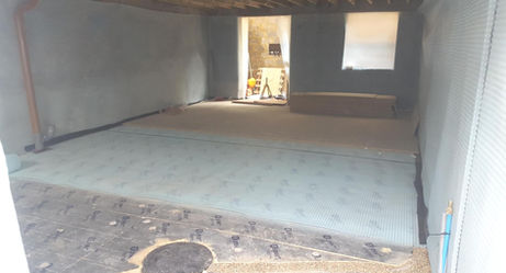 A dimly lit, unfinished basement with exposed beams and gray waterproofing membrane on walls and floor