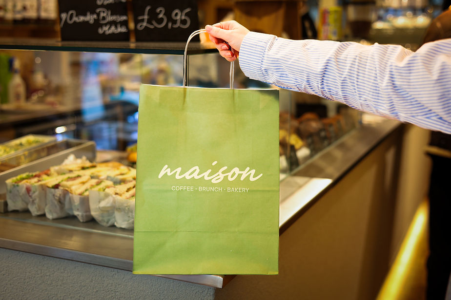 Hand holding a green Maison Coffee Brunch Bakery paper bag at a café counter