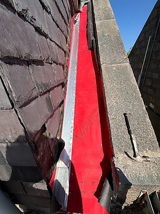 A roof valley with a vibrant red waterproof membrane runs between slate tiles and a stone wall