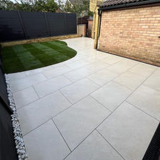 A beige and tan stone patio adjacent to a brick house, bordered by a low brick wall with niches, a wooden deck, and a white-framed conservatory, enclosed by a grey privacy fence