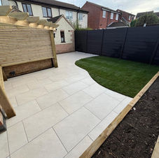 A landscaped backyard with a paved stone patio, raised garden beds, gravel borders, wheelbarrows, and a circular grass patch, enclosed by a tall wooden fence