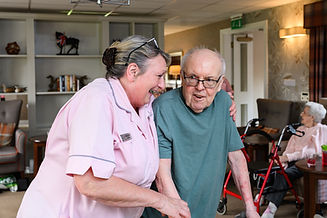 A caregiver in a pink uniform supports an elderly man using a walker