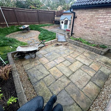 A newly installed patio with multicolored stone tiles in earthy tones, arranged in a random mosaic pattern, bordered by brick walls and fencing, with a conservatory on the side