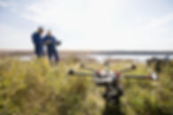 Two engineers in blue uniforms and helmets examine a tablet near a large drone in a grassy field by a lake, suggesting a calm, focused survey