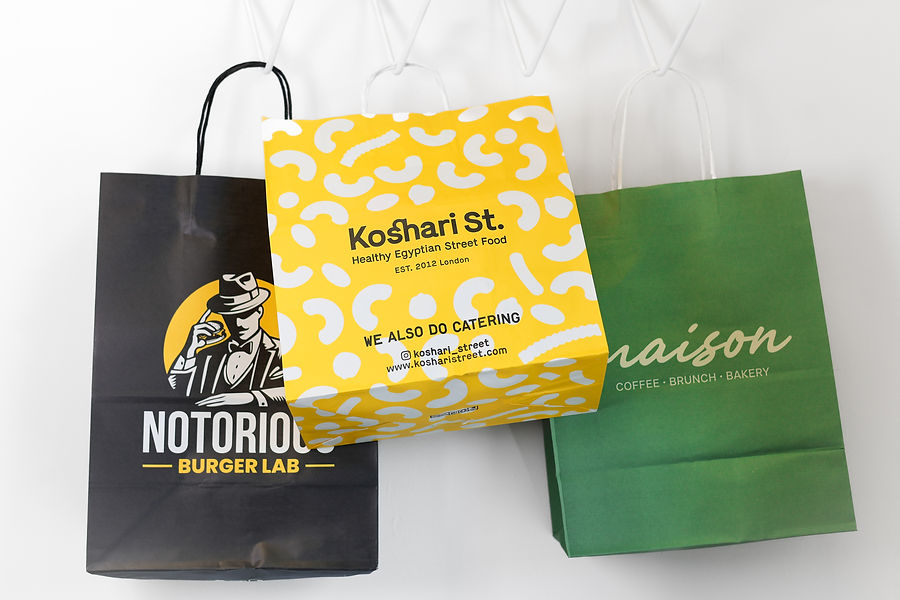 Three branded paper shopping bags from Notorious, Koshari St., and Maison hanging on a white wall