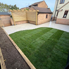 A backyard garden featuring a circular stone patio with wheelbarrows, green grass, a stone pathway, a playhouse, and a wooden shed, with a person working near the house