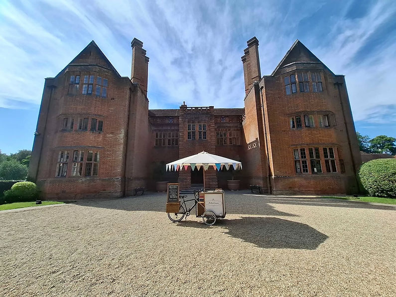 A vintage ice cream tricycle parked on a gravel driveway in front of a grand brick manor house