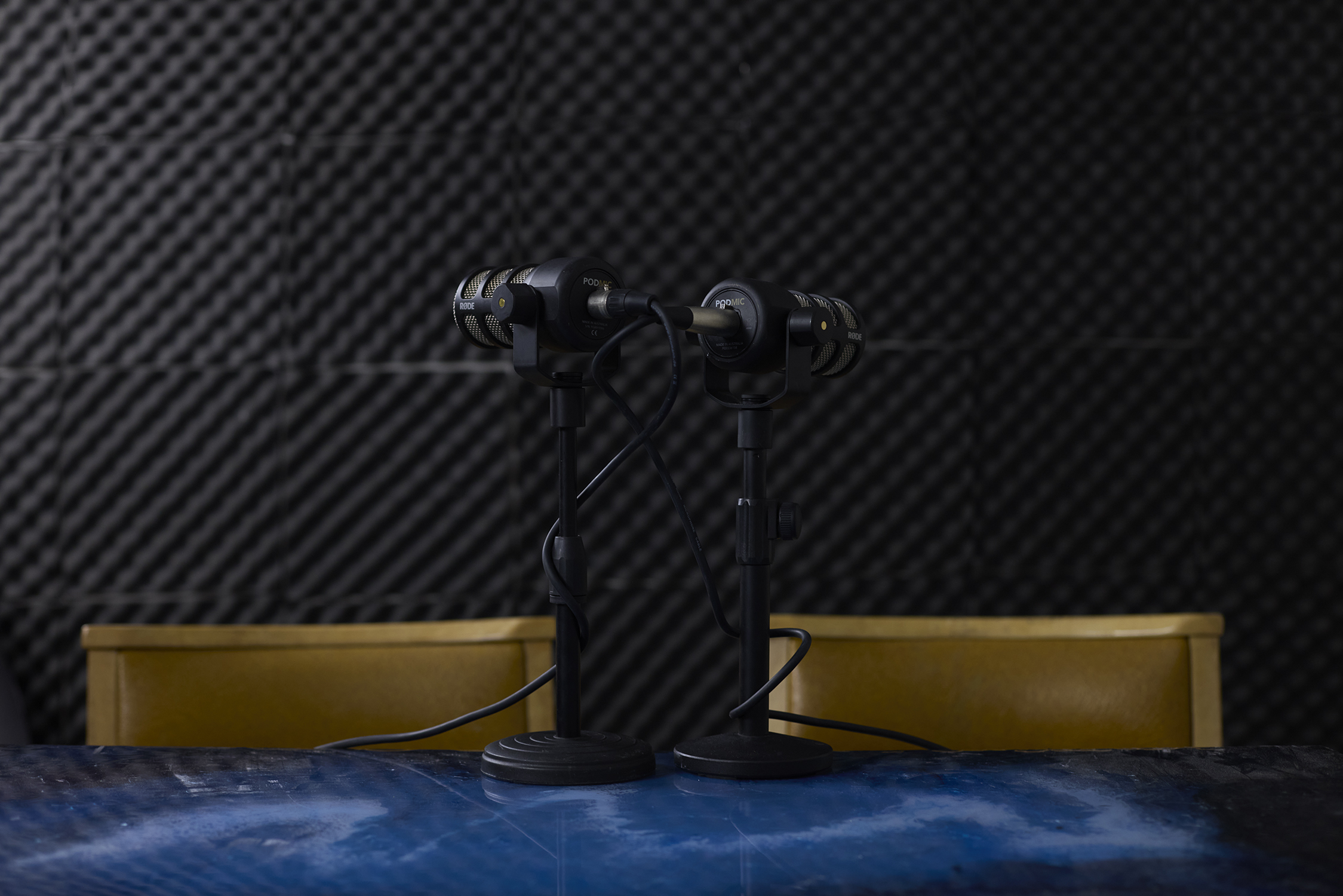 Two microphones on stands on a blue table with acoustic wall and yellow chairs in Vessel Liverpool's podcast room
