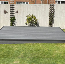 A modern backyard with a tiled patio, curved grass lawn, wooden pergola, and raised soil bed, bordered by a black wooden fence and neighboring houses