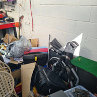 Cluttered garage interior with stacked cardboard boxes, plastic bags, tools, and miscellaneous items