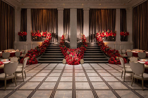 Elegant ballroom with marble floor, grand dual staircases adorned with vibrant red flowers, soft lighting