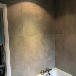 Bathroom view showing a white bathtub with chrome fixtures set against a wall of large, matte gray tiles illuminated by overhead lighting.